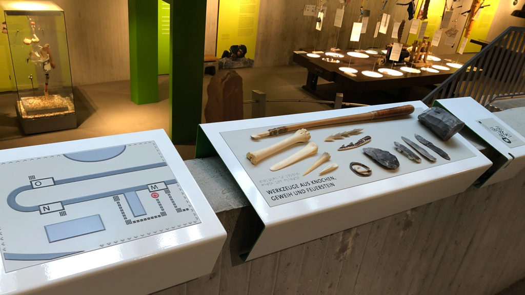 Ein Tastobjekt mit verschiedenen Werkzeugen aus Knochen, Geweih und Feuerstein. Daneben der QR-Code für Neanderthal: Memories und eine Orientierungskarte. 